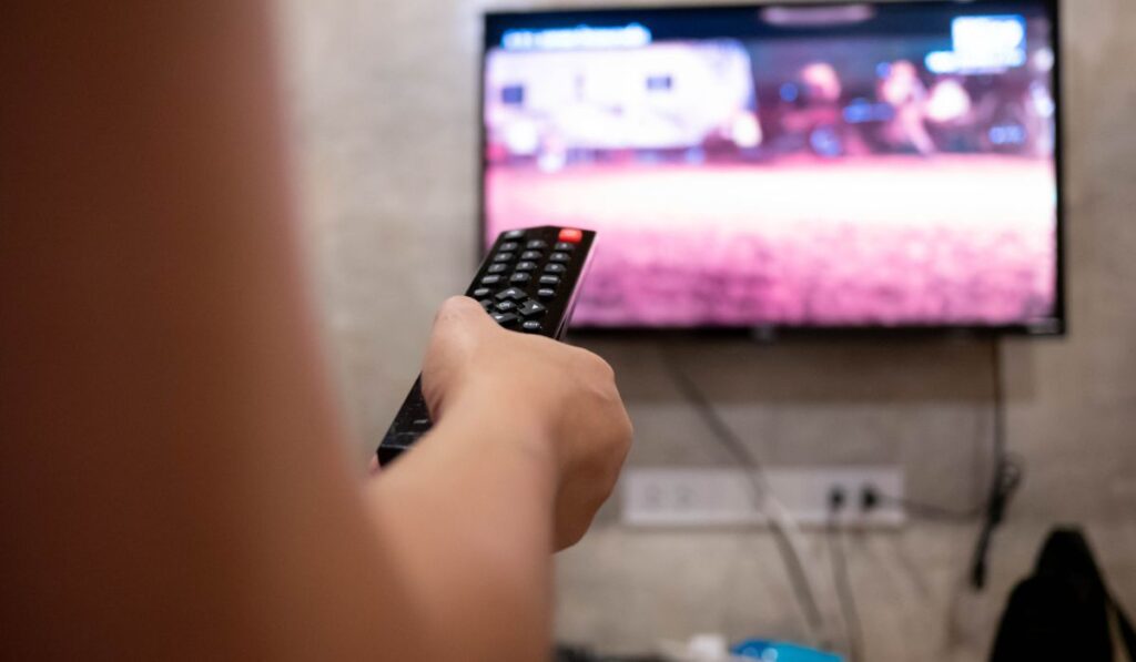 Азиатская молодая женщина смотрит телевизор в комнате с пультом дистанционного управления в руке, чтобы отрегулировать громкость