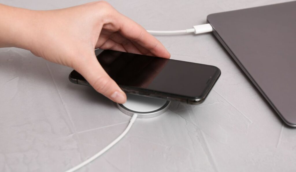 Женщина кладет смартфон на беспроводное зарядное устройство, подключенное к ноутбуку, за светло-серым столом