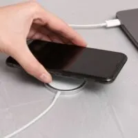 Woman putting smartphone on wireless charger connected to laptop at light grey table