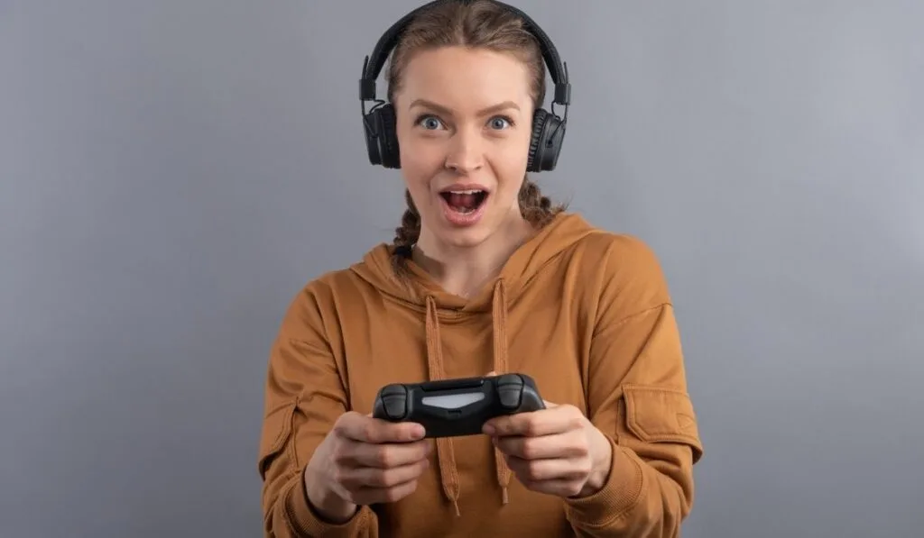Close-up view of emotional woman using joystick and headphone on gray background 