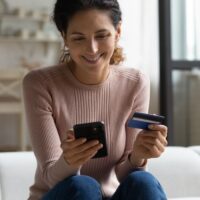 Smiling latina lady holding phone bank card make online shopping