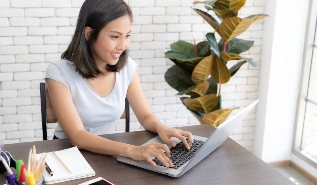 Портрет улыбающейся счастливой красивой азиатской женщины, использующей технологию портативного компьютера, сидя на столе и печатая на клавиатуре