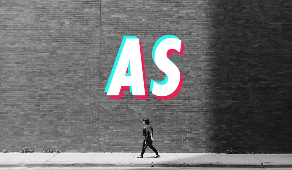 Черно-белое фото женщины, идущей рядом с кирпичным зданием, буквы «AS» показаны выше.