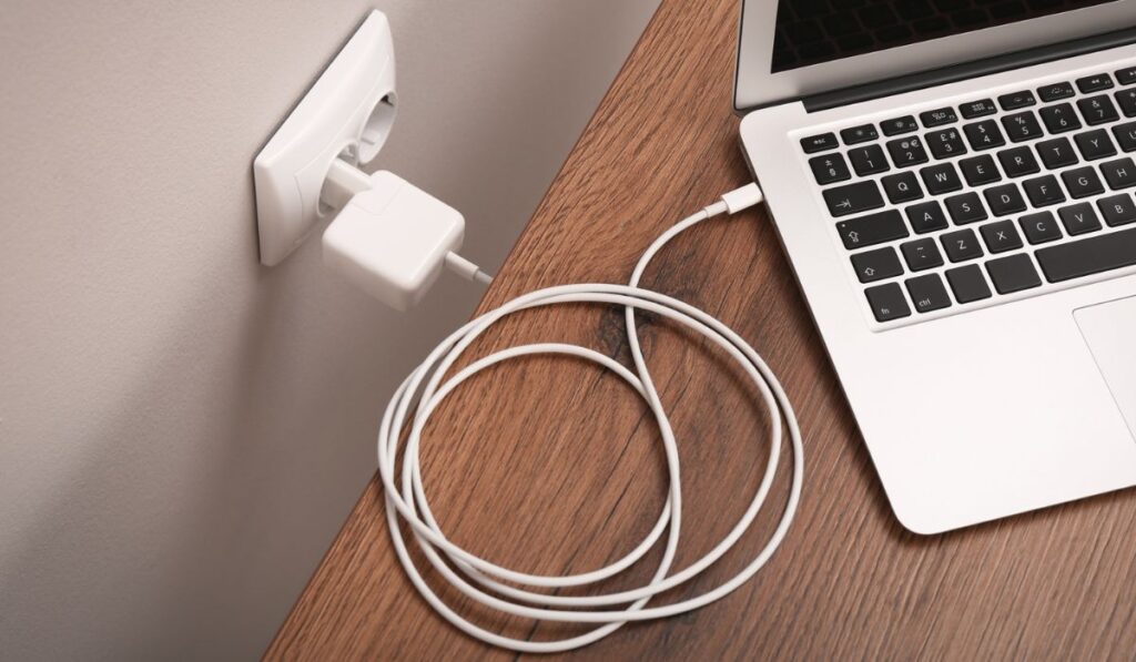 Charging laptop with power adapter in electrical socket on wooden table