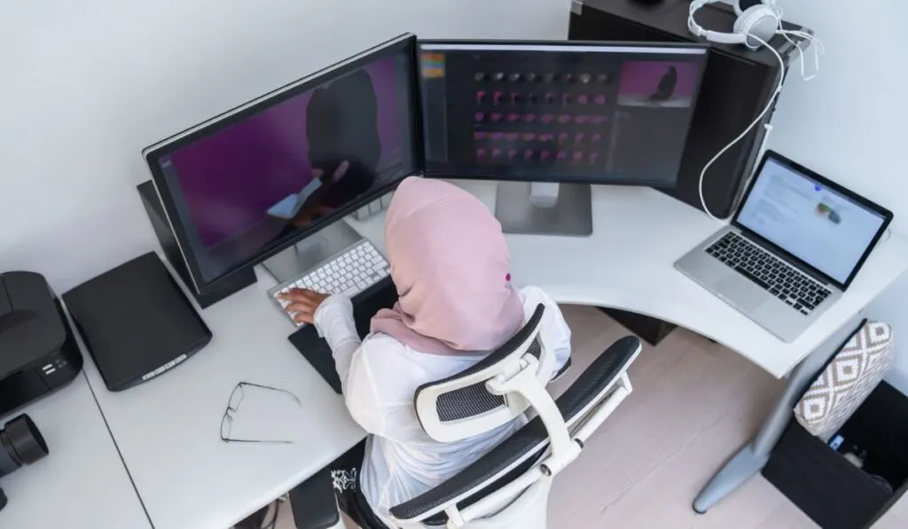 Женский арабский творческий профессионал, работающий в домашнем офисе на настольном компьютере с видом сверху на монитор с двумя экранами
